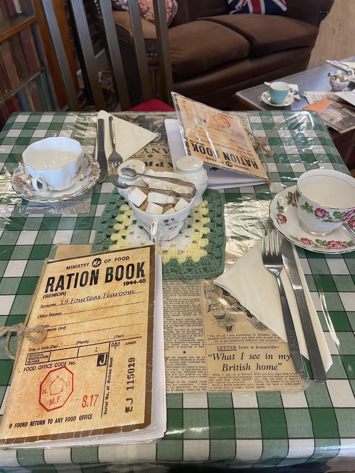 A table laid with a Ministry of Food ration book menu, floral teacups and sugar lumps on a green gingham cloth
